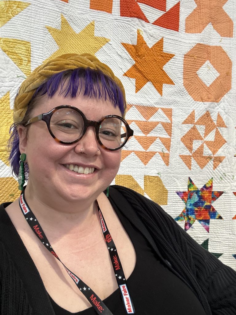 Smiling person with purple hair and glasses, wearing a lanyard, in front of a colorful quilt with geometric star patterns in orange and yellow.