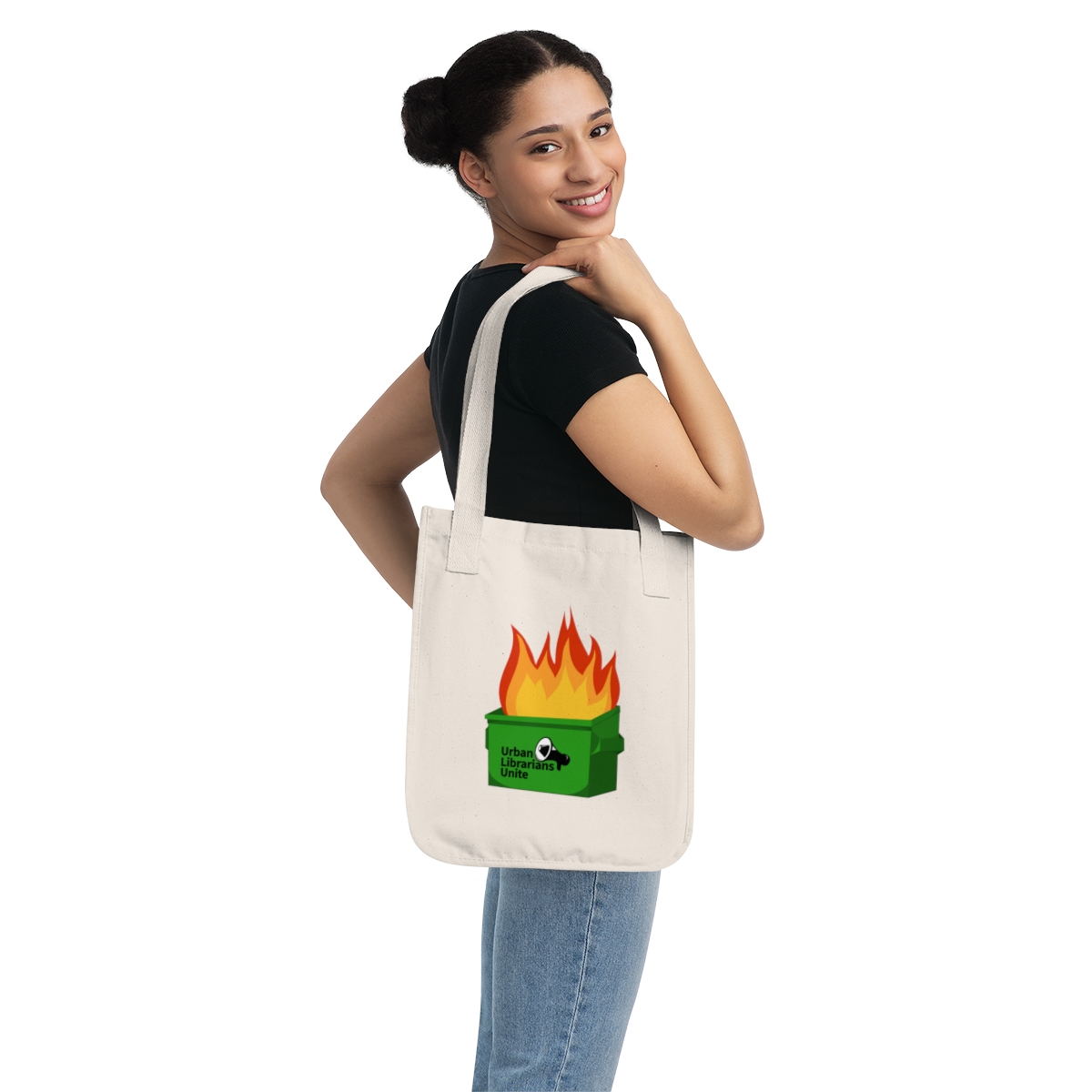A smiling woman with her hair in a bun carries a natural cream-colored canvas tote bag over her shoulder. The bag features an illustration of a green dumpster engulfed in orange and red flames, with the text "Urban Librarians Unite" and a megaphone icon on the front.