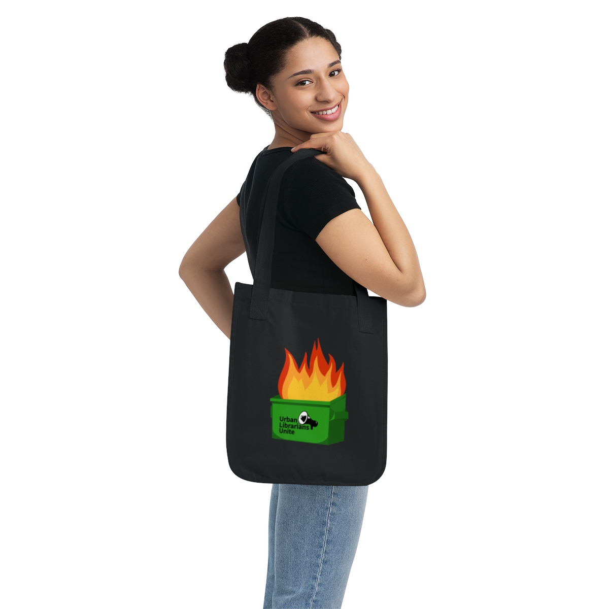 A smiling woman with her hair in a bun carries a black canvas tote bag over her shoulder. The bag features an illustration of a green dumpster engulfed in orange and red flames, with the text "Urban Librarians Unite" and a megaphone icon on the front.