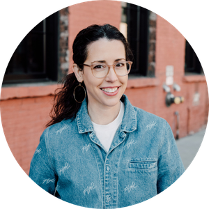 Headshot of a smiling woman with dark hair in a ponytail, wearing glasses, hoop earrings, and a denim jacket, standing outdoors.