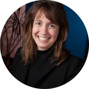 Headshot of a smiling woman with long brown hair, wearing a black turtleneck, with a dark blue background.
