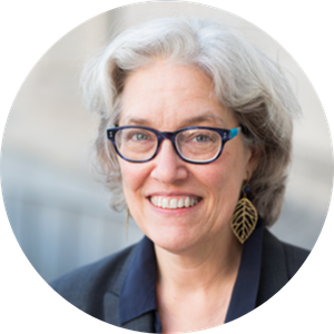 Headshot of a smiling woman with short gray hair, wearing blue-framed glasses, leaf-shaped earrings, and a navy blazer.