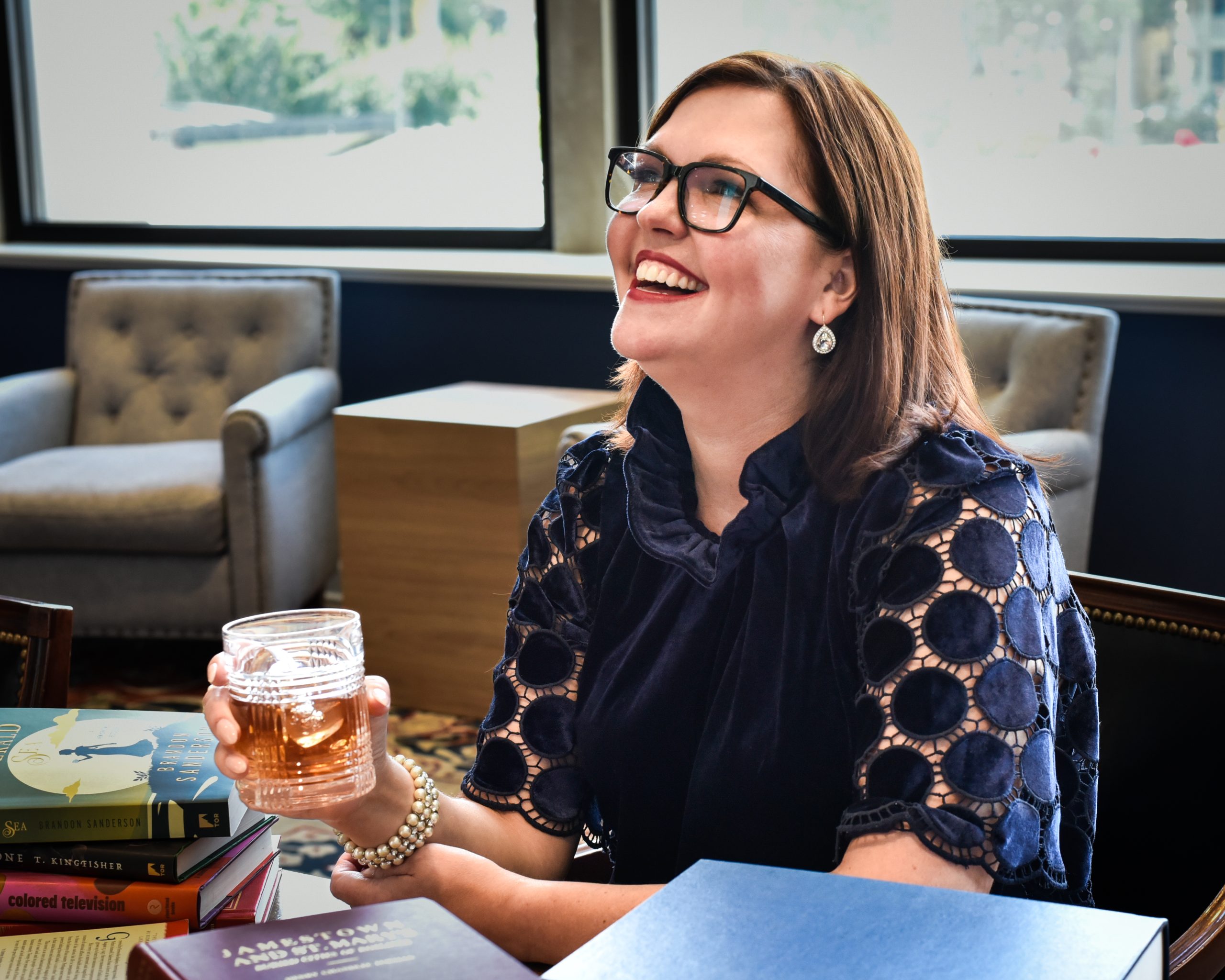 smiling woman with brown hair and glasses sitting at a table covbered in boo9ks with a glass of whiskey in her hand