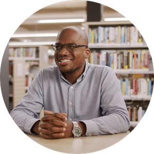 Headshot of a smiling man with a shaved head, wearing glasses and a light blue button-down shirt, seated in front of bookshelves.