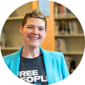 Headshot of a smiling person with short, spiked hair, wearing hoop earrings, a "Free People Read Freely" t-shirt, and a bright turquoise blazer, standing in front of bookshelves.