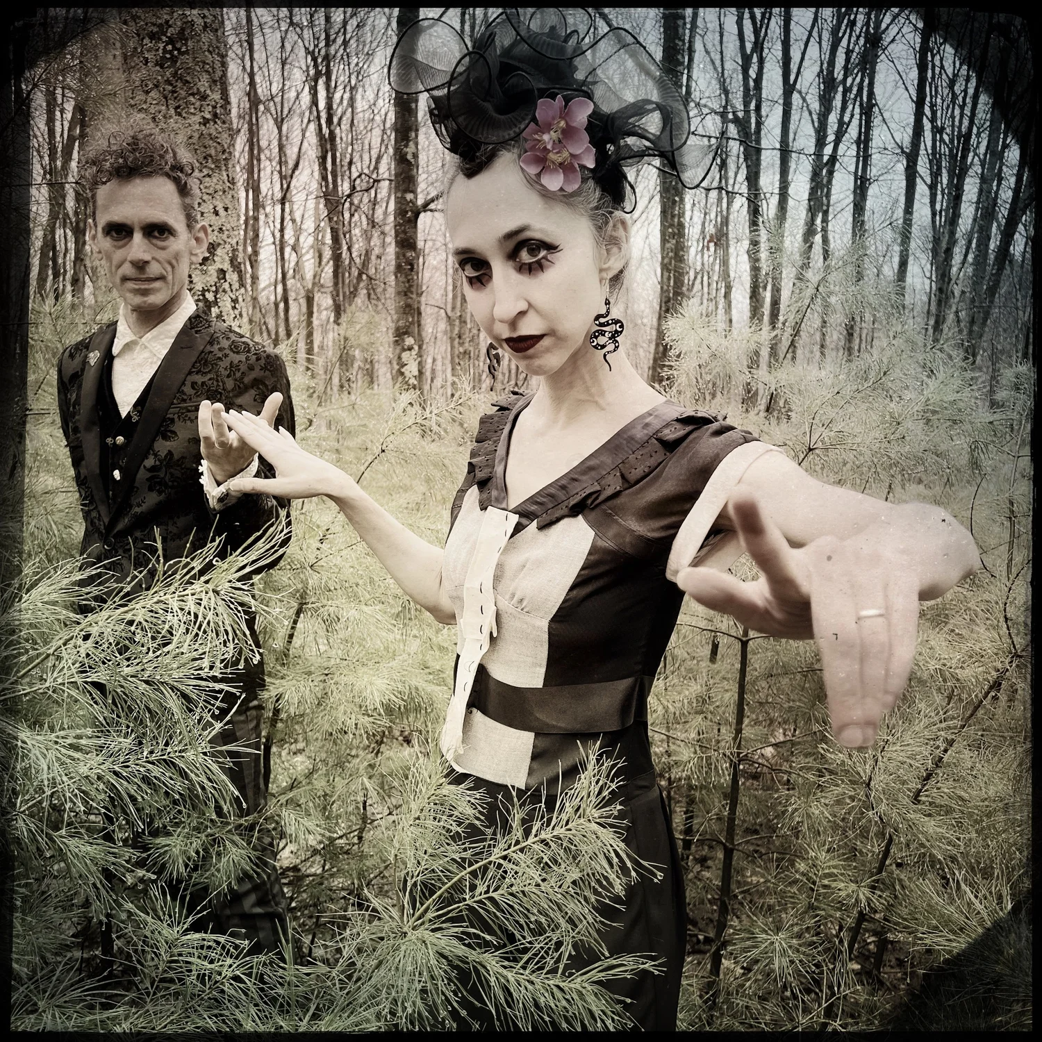 Sepia-toned photo of two people in gothic Victorian-style attire standing among pine branches in a bare winter forest. A woman in the foreground wears dark dramatic eye makeup, a floral hair piece with black netting, and a two-tone brown and cream dress, gesturing toward the camera with both hands. A man stands behind her in a dark brocade vest and jacket.