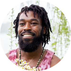 Headshot of a smiling man with long locs and a beard, wearing a pink tie-dye tank top and a floral necklace, photographed outdoors.