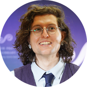 Headshot of a smiling person with curly hair and round glasses, wearing a vest, shirt, and tie, with a purple patterned background.