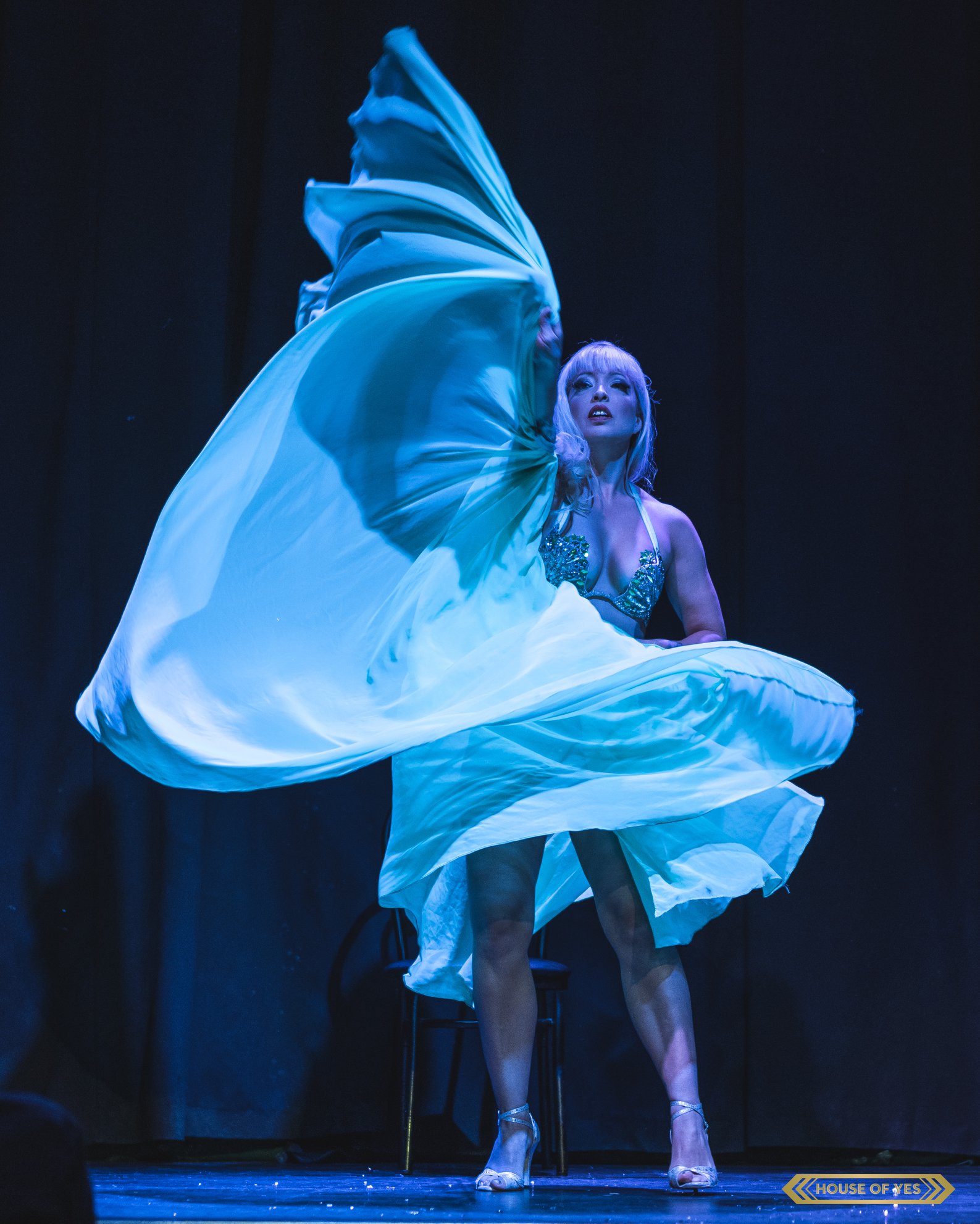 A performer on stage swirls a large flowing fan of pale blue-white silk fabric, illuminated by dramatic blue stage lighting. She wears a sequined bra top and silver heels, with platinum blonde hair, against a dark theatrical backdrop. 'House of Yes' logo in the corner.